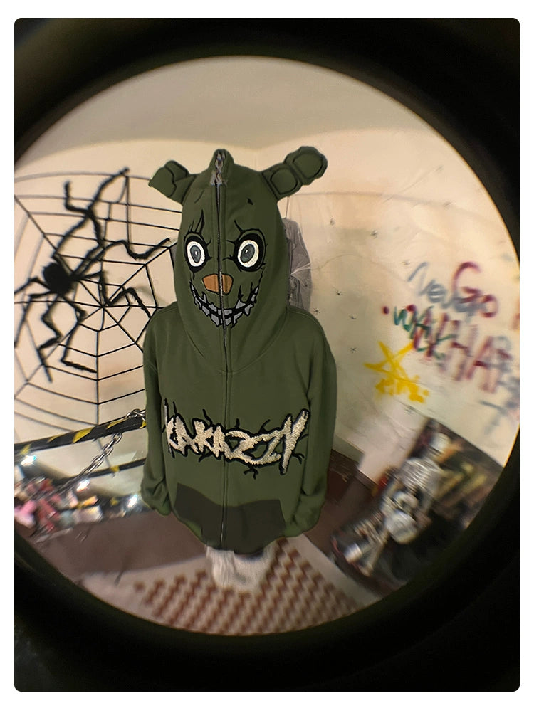 In a room decorated with wall graffiti and a large spider decoration, someone wears a Seakoff Springtrap-Inspired Hoodie. This oversized, 400GSM green zip-up sweatshirt from Five Nights at Freddy&