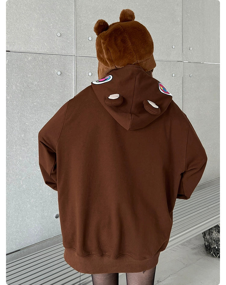 A person models the Seakoff Cute Bear Hoodie, a charming 400GSM oversized brown zip-up streetwear sweatshirt with bear ears on the hood. Small eyes and ears adorn the back, creating an adorable look against a tiled wall and bench backdrop.