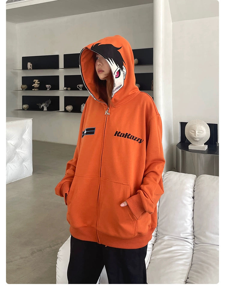 Indoors, a person dons the Seakoff Fire Force Inspired Orange Zip-Up Hoodie, an oversized 400GSM anime streetwear piece with hood up. It features a logo and text on the front. Behind them, a modern shelf with decorative items and a sculpted head stands amidst neutral room tones.