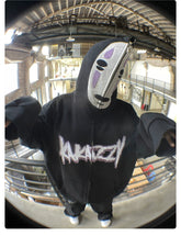 Someone indoors is wearing a Spirited Away No-Face Anime Hoodie, an oversized 400GSM zip-up sweatshirt with "Krazy" on it. Their face is hidden by a white oval mask with black and purple markings, set against an industrial backdrop ideal for anime fans.