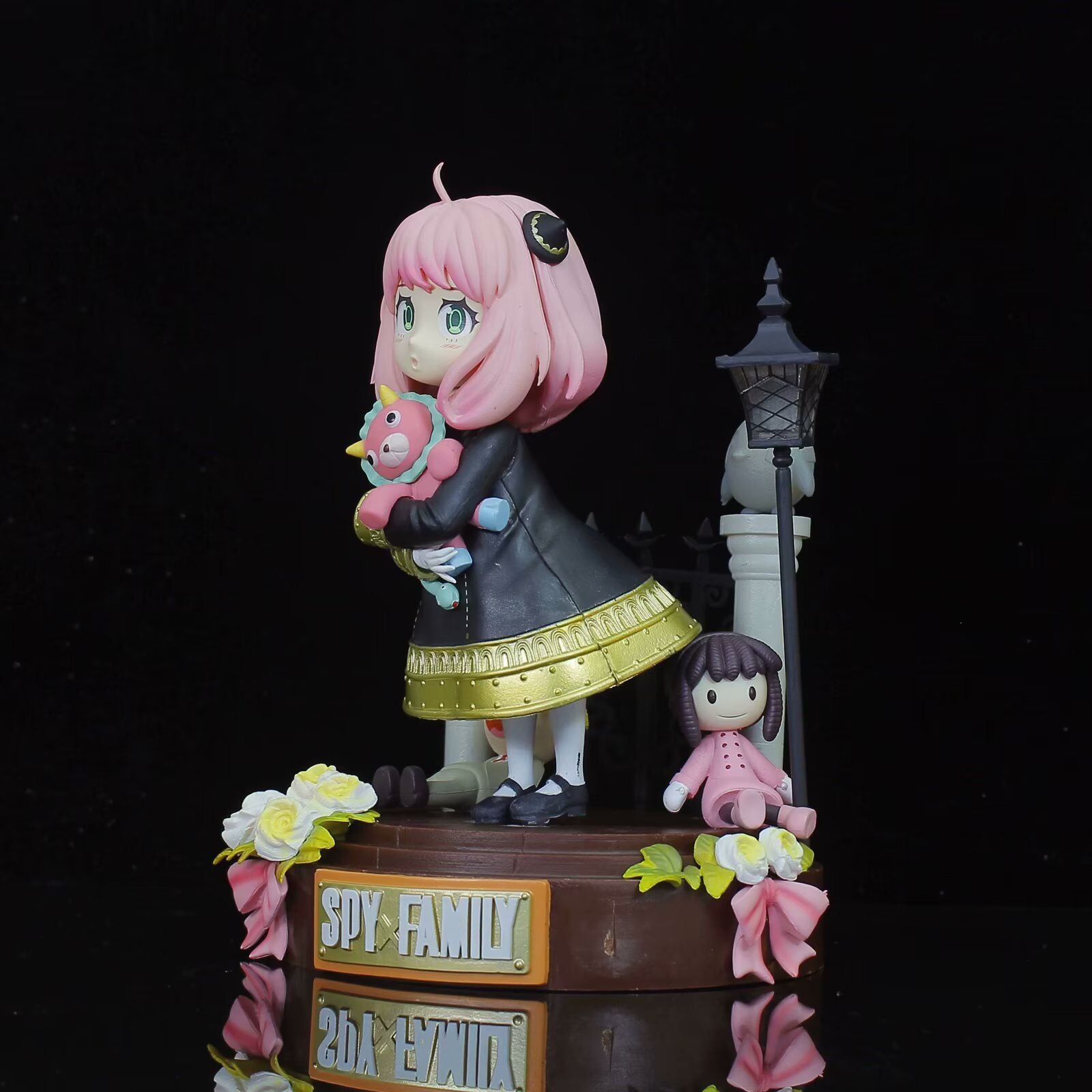 The Spy × Family Anya Forger PVC Figure depicts a young girl with pink hair holding a plush toy, dressed in a black and gold outfit on a flower-adorned base labeled "Spy Family," featuring a doll and vintage lamp post.