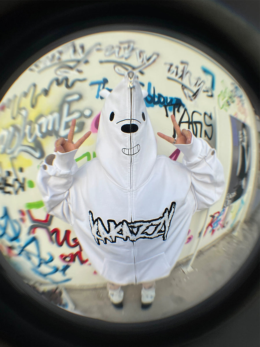 Against a vibrant graffiti wall, a person models the Cute White Bear Hoodie by Seakoff, offering an oversized fit with black embroidery. Flashing peace signs from both hands, "ANARCA" stands out on the hoodie. The scene is creatively captured with a fisheye lens effect.