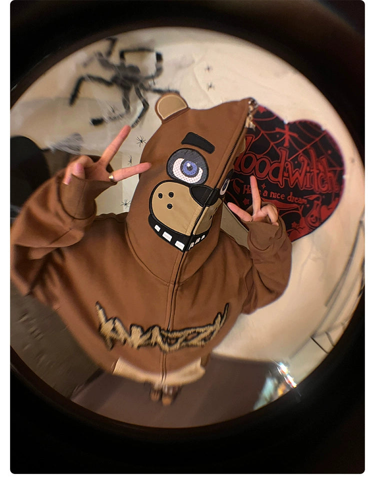 A person in a Seakoff Freddy-Inspired Hoodie, an oversized brown zip-up sweatshirt with a bear face design, has the hood up and makes peace signs. The background includes a spider decoration and "Goodwitch" sign, ideal for Five Nights at Freddy&