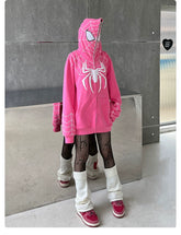 A superhero fan wears a Seakoff Spider-Inspired Hoodie, a bright pink oversized zip-up sweatshirt in cozy 400GSM cotton with white web designs. Paired with black lace leggings, white leg warmers, and pink sneakers, their face is hidden by the hood against a metallic indoor backdrop.