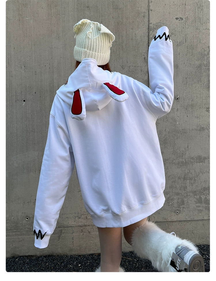 A person in a Seakoff Fierce Bunny Hoodie, featuring cartoon eyes and red rabbit ears, poses against a concrete wall. The oversized fit is complemented by a white knit hat and fluffy shoes, as they pose with one arm raised.