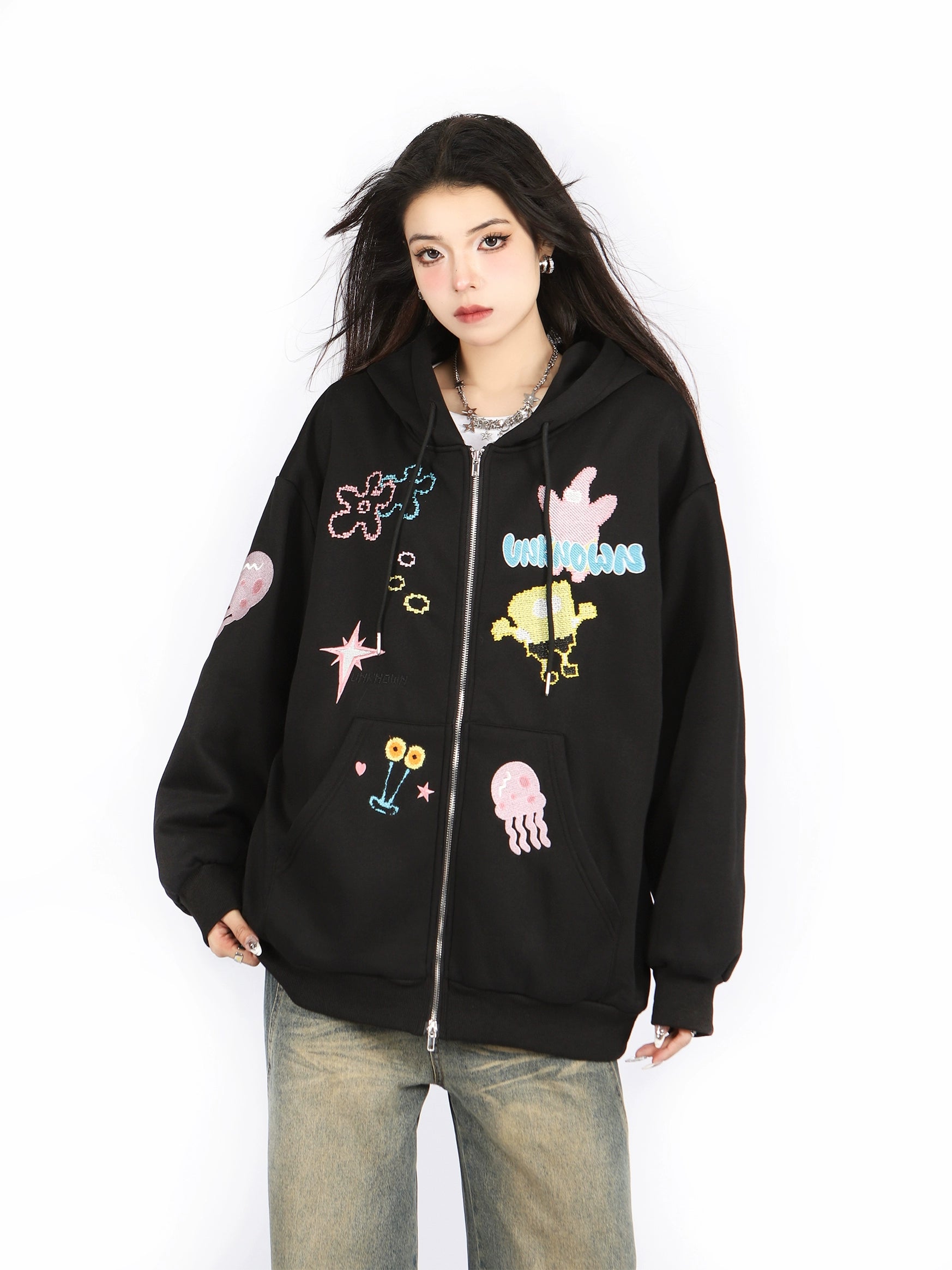 A person with long hair wears a Seakoff Kawaii Pastel Cartoon Hoodie featuring playful space-themed embroidered designs, including stars and planets. Pairing it with baggy jeans, they have a neutral expression and stand against a plain white background.