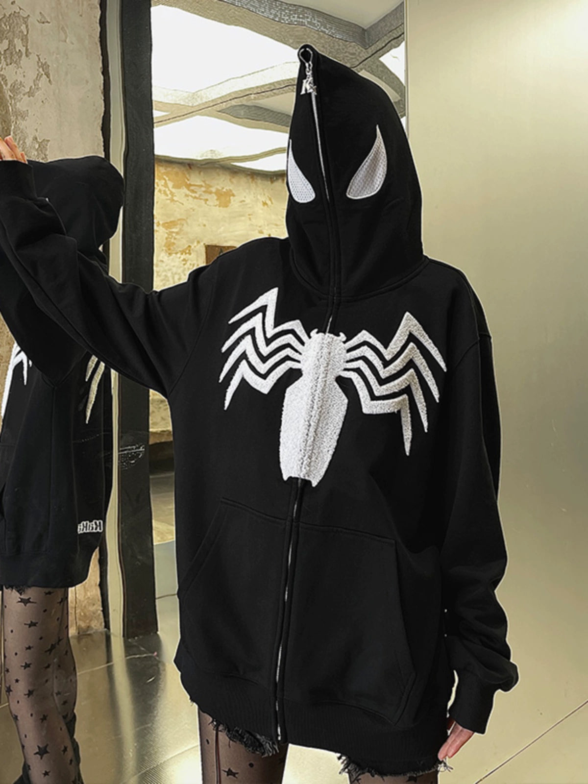 A person stands indoors in a Seakoff Venom Spider Hoodie, a black and white 400GSM oversized zip-up sweatshirt with a distinctive spider emblem. The hood zipped up adds an air of mystery. In the reflection, eye-catching patterned leggings complete this streetwear fan&