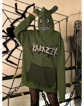 A person in a Seakoff Springtrap-Inspired Hoodie—a green oversized zip-up with 400GSM fabric—stands by Halloween decor featuring a large spider and skeletal figure. The hoodie, ideal for Five Nights at Freddy&