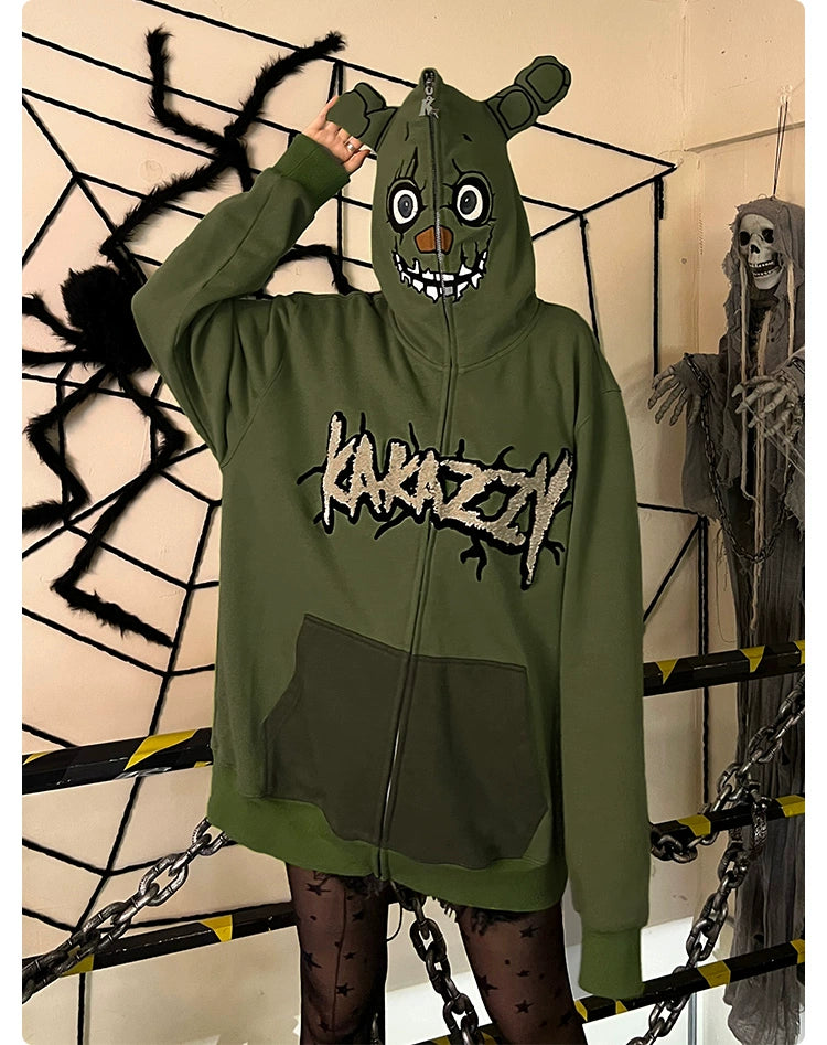 A person in a Seakoff Springtrap-Inspired Hoodie—a green oversized zip-up with 400GSM fabric—stands by Halloween decor featuring a large spider and skeletal figure. The hoodie, ideal for Five Nights at Freddy&