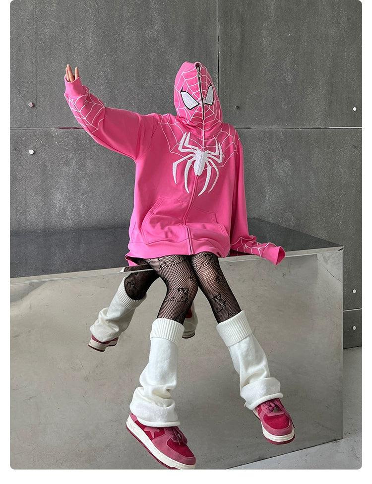 A person poses on a reflective surface in a Seakoff Spider-Inspired Hoodie, an oversized 400GSM pink zip-up sweatshirt with web-designed hood and spider logo. They pair it with patterned tights, leg warmers, and pink sneakers—a superhero fan&