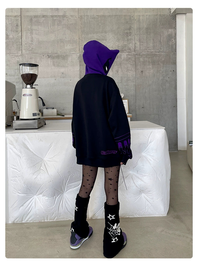 A person is seen from behind, wearing a Seakoff Dark Hero Hoodie—an oversized black and purple superhero zip-up sweatshirt. They pair it with sheer black star-patterned tights, starry black boots, and purple sneakers in a modern room with concrete walls and a coffee grinder.
