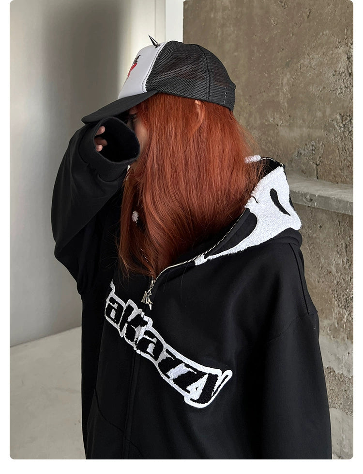 A person with long red hair, wearing a Seakoff Scream-Inspired Hoodie—a 400GSM oversized black zip-up horror movie sweatshirt—and a black-and-white cap, subtly hides part of their face with their sleeve. Against a beige wall, they exude a mysterious aura that horror fans might find intriguing.