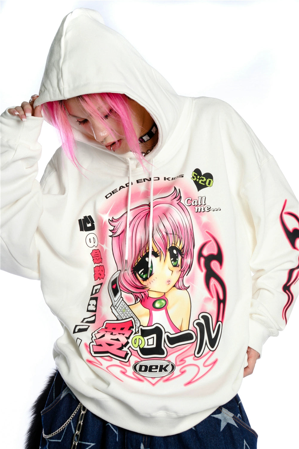 A person with pink hair and kawaii style adjusts the hood of their white Seakoff hoodie, adorned with vibrant anime artwork of large-eyed characters and Japanese text, capturing the nostalgic charm of Y2K aesthetic.