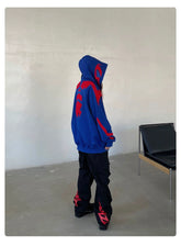 A person in a Seakoff Superhero Spider Hoodie, an oversized blue and red zip-up sweatshirt, stands in a minimalist room wearing black pants with red designs and black shoes. In the background is a black chair with a wooden frame, against plain light-colored walls and floor.