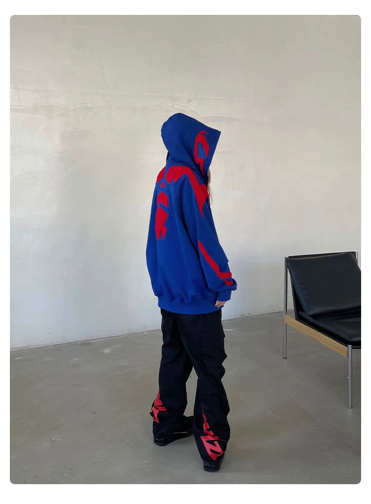 A person in a Seakoff Superhero Spider Hoodie, an oversized blue and red zip-up sweatshirt, stands in a minimalist room wearing black pants with red designs and black shoes. In the background is a black chair with a wooden frame, against plain light-colored walls and floor.