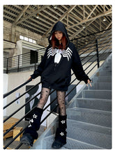 A person in a Seakoff Venom Spider Hoodie—an oversized black and white zip-up sweatshirt—paired with black star-patterned tights and leg warmers, stands on an industrial-style staircase indoors, crafting a striking look sure to captivate streetwear enthusiasts.