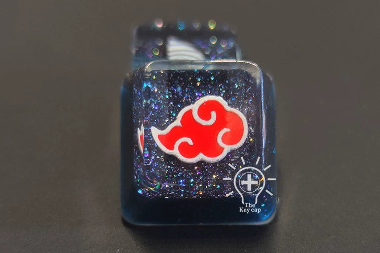 A close-up of a resin keycap from the Naruto Symbols Keycap Set showcases a red cloud design on a dark, glittery background. The glossy, illuminated finish features a small cross logo with "The Keycap" text.