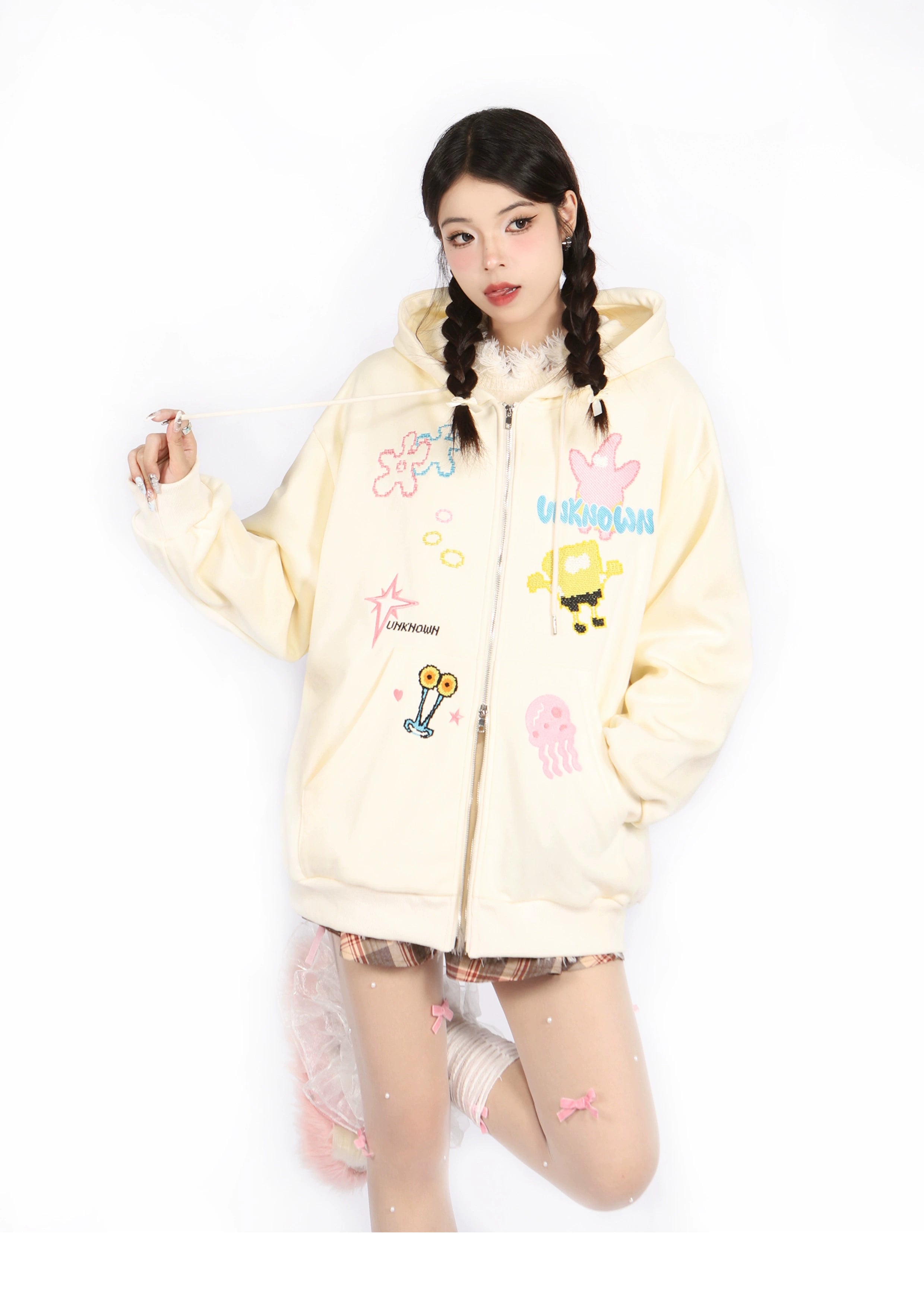 A person with braided hair, embodying anime fashion, wears Seakoff’s Kawaii Pastel Cartoon Hoodie featuring playful embroidered designs. They pair it with plaid shorts and knee-high socks with bows, against a white background.