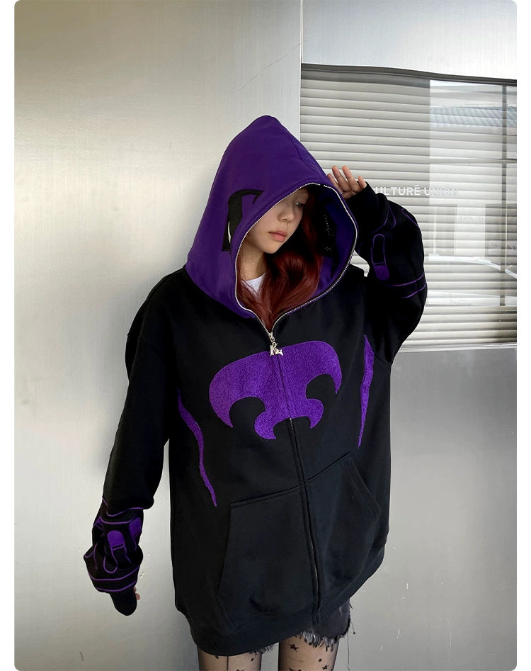 Against a metallic wall, a person wears the Seakoff Dark Hero Hoodie. The black and purple oversized zip-up partially covers their face with its distinctive pattern, evoking a dark hero vibe. Long hair cascades under the hood of this 400GSM streetwear sweatshirt.
