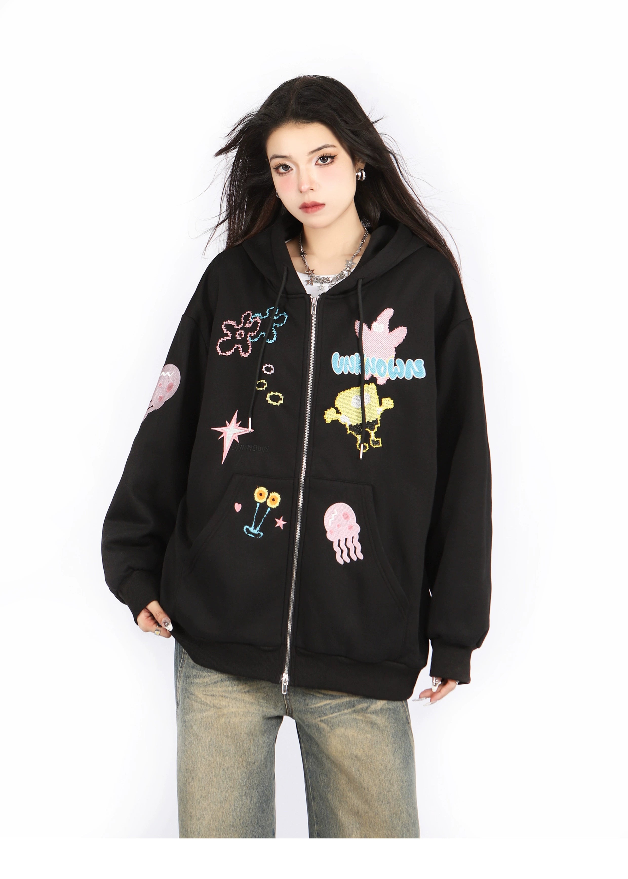A person with long dark hair wears the Seakoff Kawaii Pastel Cartoon Hoodie, a black full-zip with playful embroidered anime-inspired designs, and light jeans against a plain white background.