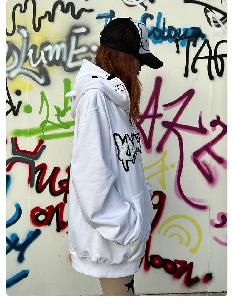 A person in a Seakoff Cute White Bear Hoodie—400GSM with a polar bear design and black embroidery—stands against graffiti. Facing sideways, their hands are tucked into the pockets, and they wear a contrasting black-and-white cap.