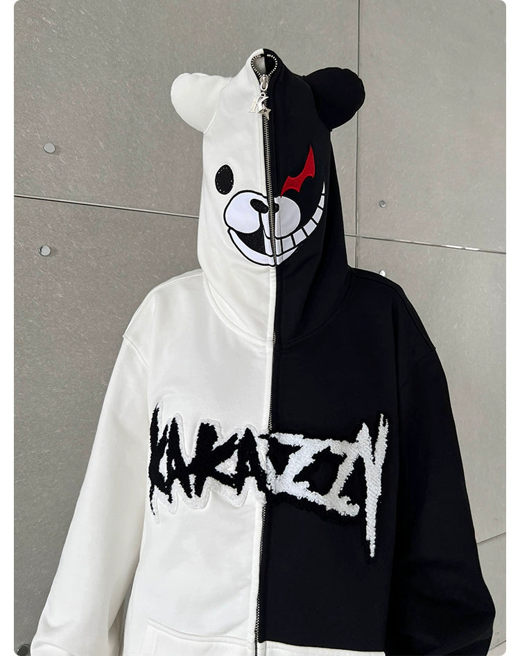 A person wears the Seakoff Monokuma-Inspired Hoodie, an oversized black and white zip-up sweatshirt featuring a bear face hood design, ideal for Danganronpa fans. The zip-up front splits into distinct halves with "Kakazy" boldly printed across it.