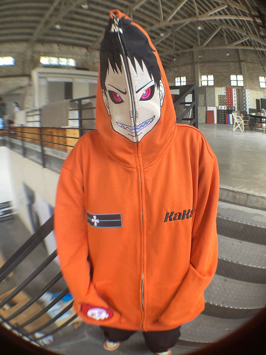 In an industrial-style space, someone sports the Seakoff Fire Force Inspired Orange Zip-Up Hoodie. This oversized anime streetwear piece features a face-covering zipper with a character print showcasing red eyes and an open mouth, capturing bold fashion vibes.