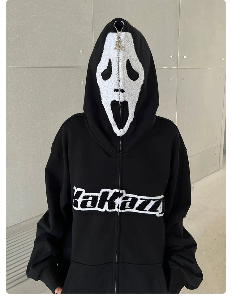 Someone wears a Seakoff hoodie featuring a white Ghostface mask design zipped up over their face, creating an eerie vibe perfect for horror fans. The bold "Karakaz" logo contrasts against the plain gray background. Product: Scream-Inspired 400GSM Oversized Black Zip-Up Horror Movie Sweatshirt.