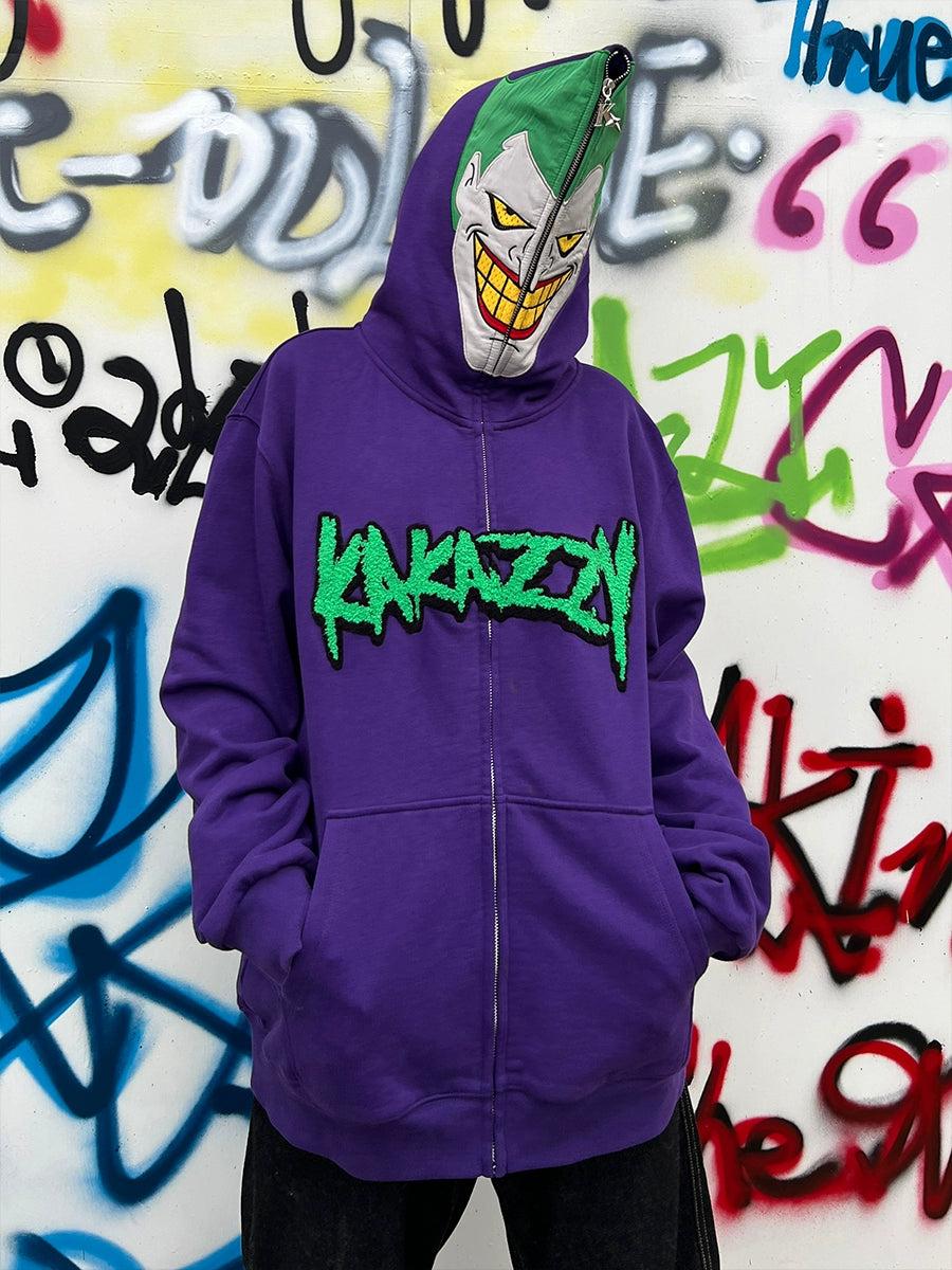 A person stands before a graffiti wall in a Seakoff Joker-Inspired Hoodie, featuring "KANAZZY" and paired with a green and white grinning mask. This oversized purple zip-up sweatshirt is perfect for supervillain fans seeking style and comfort in 400GSM cotton.