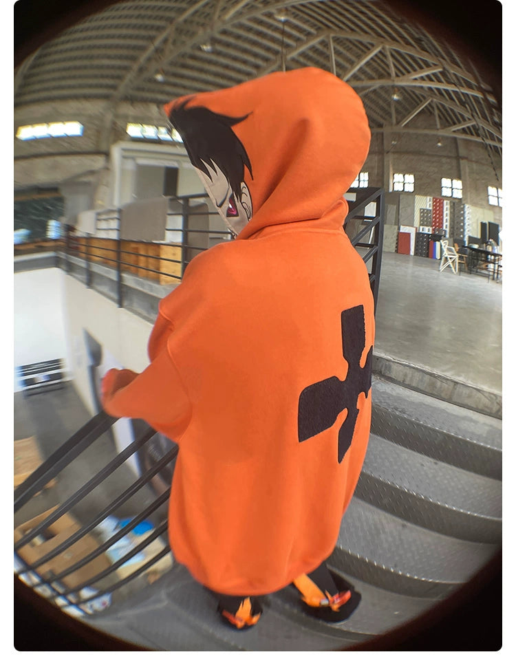 In a spacious industrial-style building, someone in a Seakoff Fire Force Inspired Orange Zip-Up Hoodie - 400GSM Oversized Anime Streetwear stands on a metal staircase. Their face is partly hidden by the hood with cartoonish anime artwork and a large black symbol on the back.