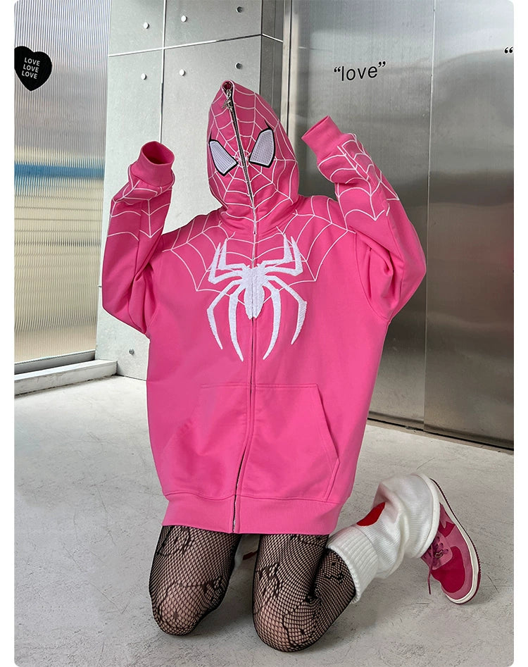 Kneeling on the floor, someone wears a Seakoff Spider-Inspired Hoodie—an oversized pink zip-up superhero sweatshirt made of plush 400GSM cotton. Their hood is up, obscuring their face. Black fishnet tights, white socks, and pink slippers complete the look against metal panels with "love" etched on the wall.