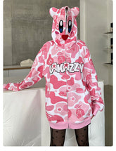 A person is wearing a Seakoff Pink Kirby-Inspired Hoodie made from 400GSM cotton with camo design, animated character motifs, and a "KAKAZY" patch. The hoodie features 3D ears on its bear-shaped hood against a modern minimalist backdrop.