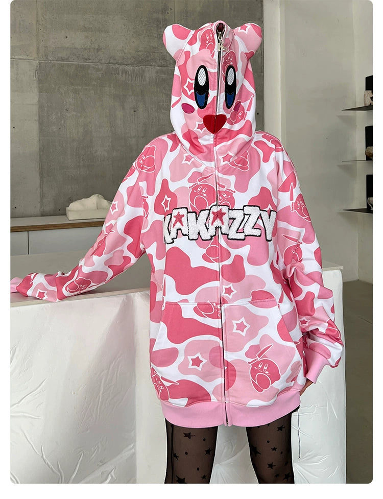 A person is wearing a Seakoff Pink Kirby-Inspired Hoodie made from 400GSM cotton with camo design, animated character motifs, and a "KAKAZY" patch. The hoodie features 3D ears on its bear-shaped hood against a modern minimalist backdrop.
