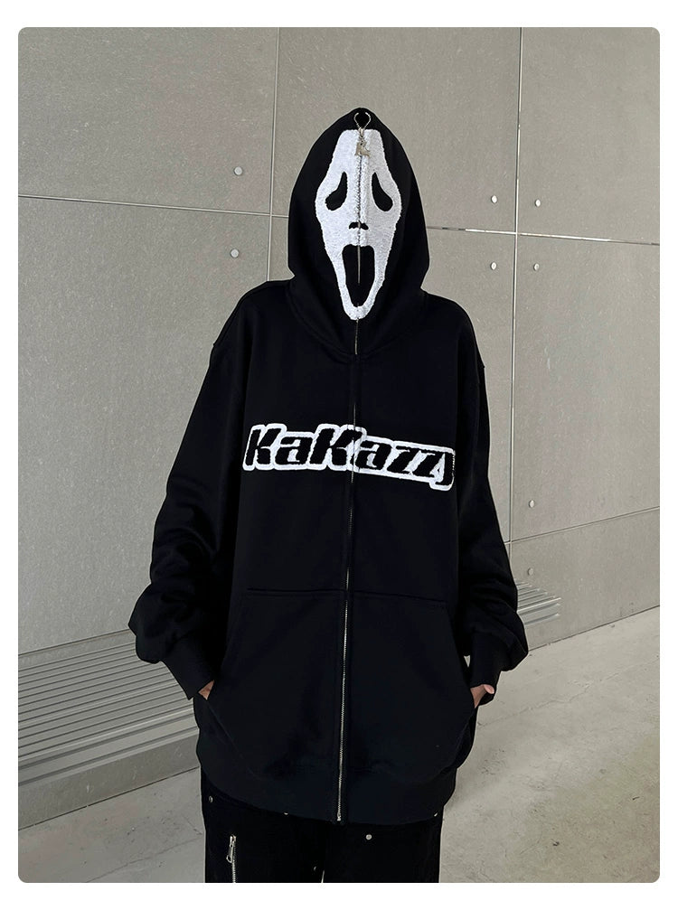 A person is wearing a Seakoff Scream-Inspired Hoodie; a 400GSM oversized black zip-up horror movie sweatshirt with "Kakaza" printed on it, featuring a hood that extends into a white Ghostface mask. Perfect for horror fans, the setting is a plain gray wall with a bench.