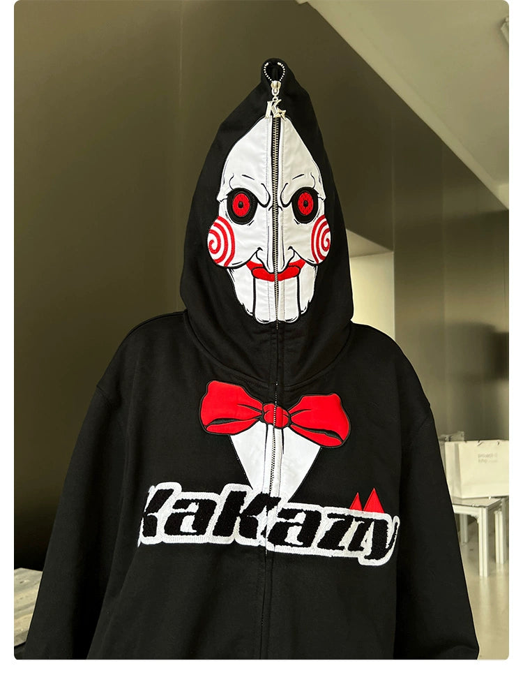 A person wears the Seakoff Saw-Inspired Hoodie, an oversized black zip-up with a mask design of Billy the Puppet from "Saw" on the hood. It features a red bow tie graphic and “Kakazy” text on the front, ideal for horror fans. The setting is a neutral indoor environment.
