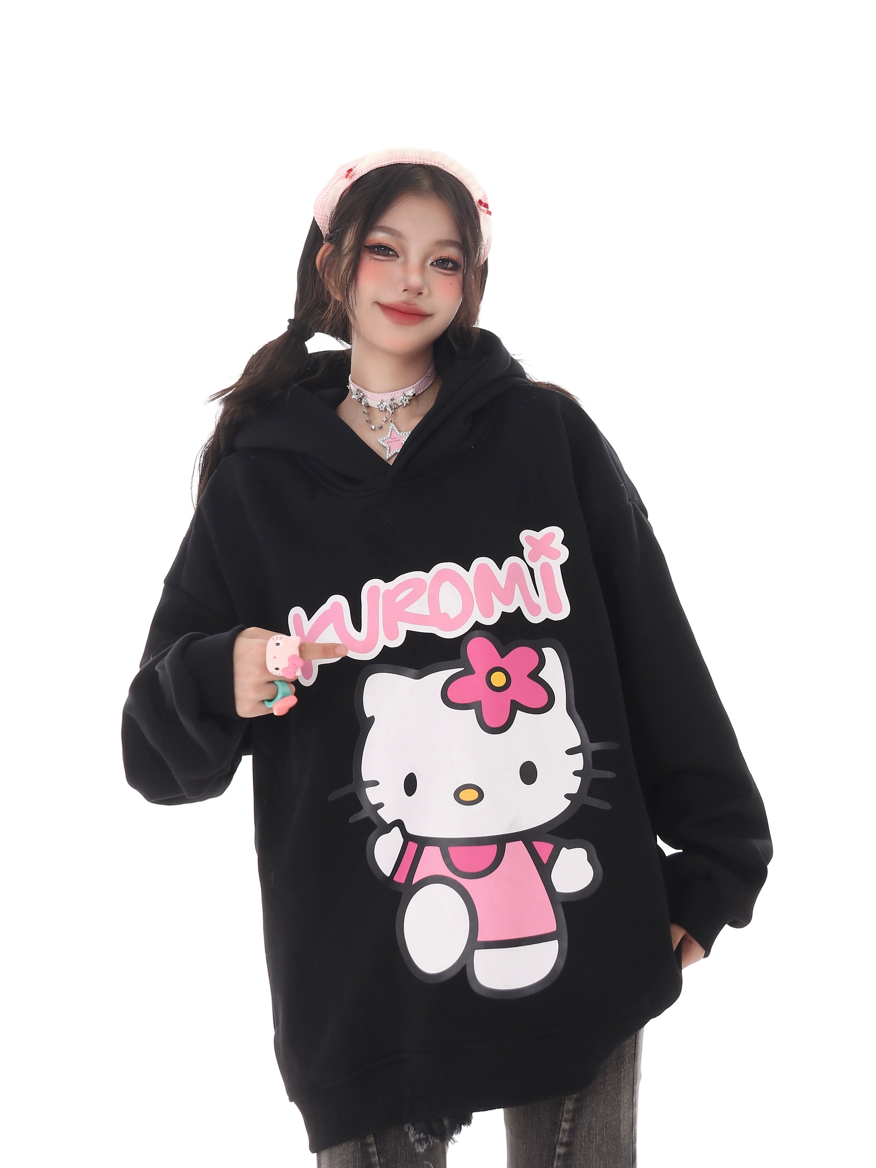 A person wears the Seakoff Kawaii Hello Kitty Oversized Hoodie, featuring a large print of Hello Kitty in a pink outfit. They hold a “Kuromi” sign and sport a pink headband against a white backdrop, embodying kawaii fashion.