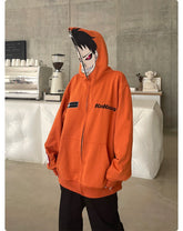 Posing by a concrete wall and coffee machine, this individual wears a Seakoff "Fire Force" Inspired Orange Zip-Up Hoodie. The oversized anime streetwear features writing, stripes, and a partially zipped character face on the hood against a white counter.
