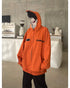 Posing by a concrete wall and coffee machine, this individual wears a Seakoff "Fire Force" Inspired Orange Zip-Up Hoodie. The oversized anime streetwear features writing, stripes, and a partially zipped character face on the hood against a white counter.