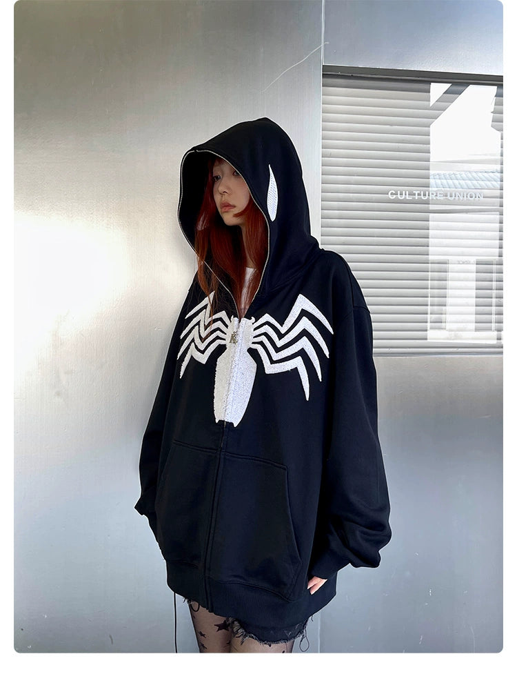 Someone in a Seakoff Venom Spider Hoodie stands indoors. The oversized black and white zip-up sweatshirt, featuring a large white spider emblem, conceals their hair with the hood up. They&