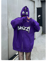 A person wears the Seakoff Fun Cartoon Face Hoodie - a purple, oversized 400GSM zip-up streetwear sweatshirt with stylized graffiti. The hood covers their face with cartoon eyes and a mouth as they stand outdoors forming a heart shape with their hands.