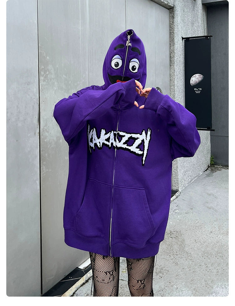 A person wears the Seakoff Fun Cartoon Face Hoodie - a purple, oversized 400GSM zip-up streetwear sweatshirt with stylized graffiti. The hood covers their face with cartoon eyes and a mouth as they stand outdoors forming a heart shape with their hands.