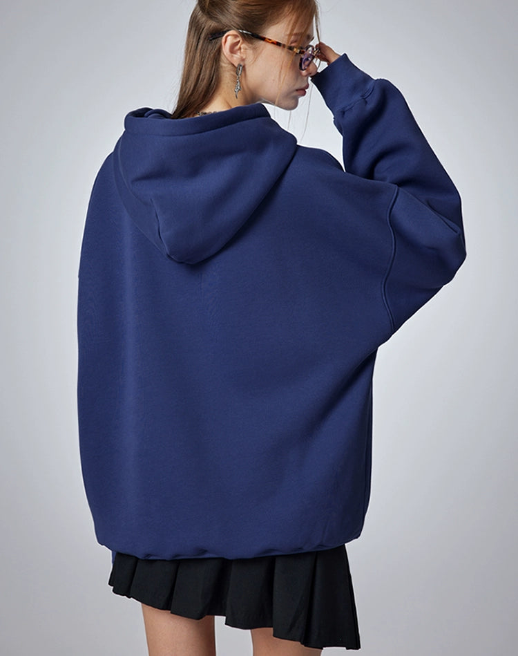 A person with long hair, seen from behind, wears a Seakoff "Oh No! I Don’t Want to Work!" oversized hoodie with a lazy cat design and a black skirt. They adjust their glasses with one hand near their face against a neutral gray background, enhancing the casual streetwear vibe.