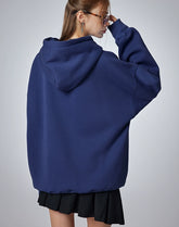 A person with long hair, seen from behind, wears a Seakoff "Oh No! I Don’t Want to Work!" oversized hoodie with a lazy cat design and a black skirt. They adjust their glasses with one hand near their face against a neutral gray background, enhancing the casual streetwear vibe.