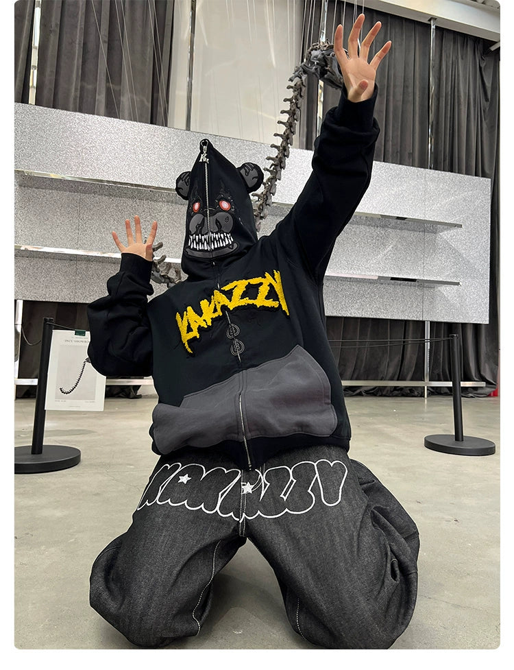 A person kneels on the floor in a Seakoff Dark Bear Demon Hoodie, crafted from 400GSM cotton with yellow embroidery, paired with a bear mask featuring teeth and red eyes. Their arms are raised, while a dinosaur skeleton tail adds to the horror ambiance.