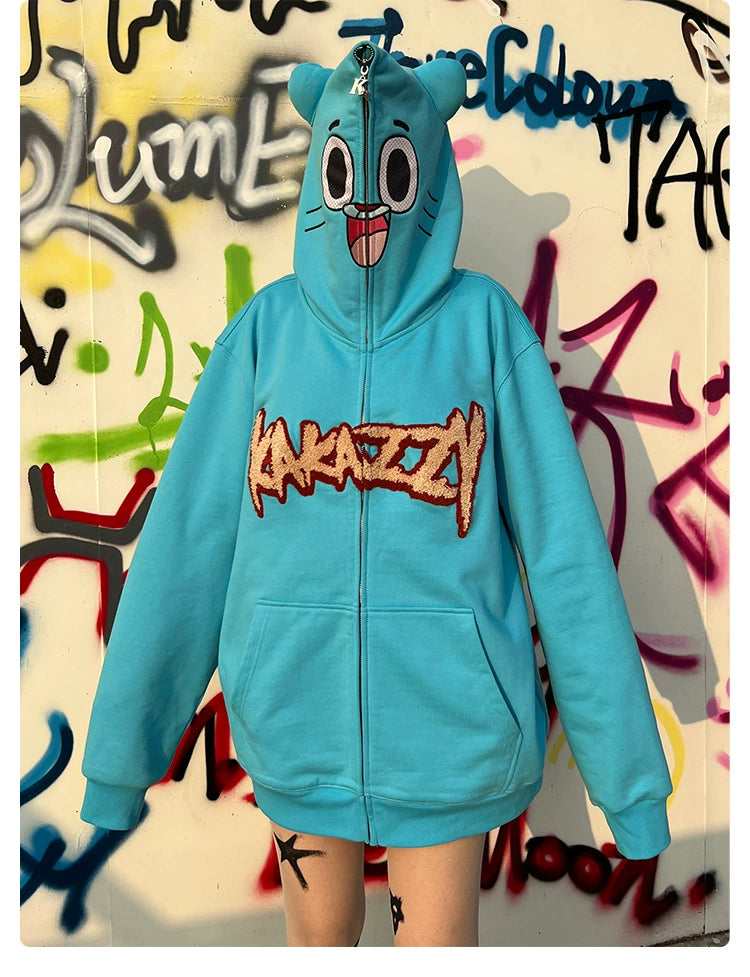 An individual sports an Adorable Blue Cartoon Hoodie by Seakoff, crafted from plush 400GSM cotton. This oversized sweatshirt features a playful face and blue hood with ears, prominently displaying the "Kanazzy" logo against a vibrant graffiti wall backdrop.