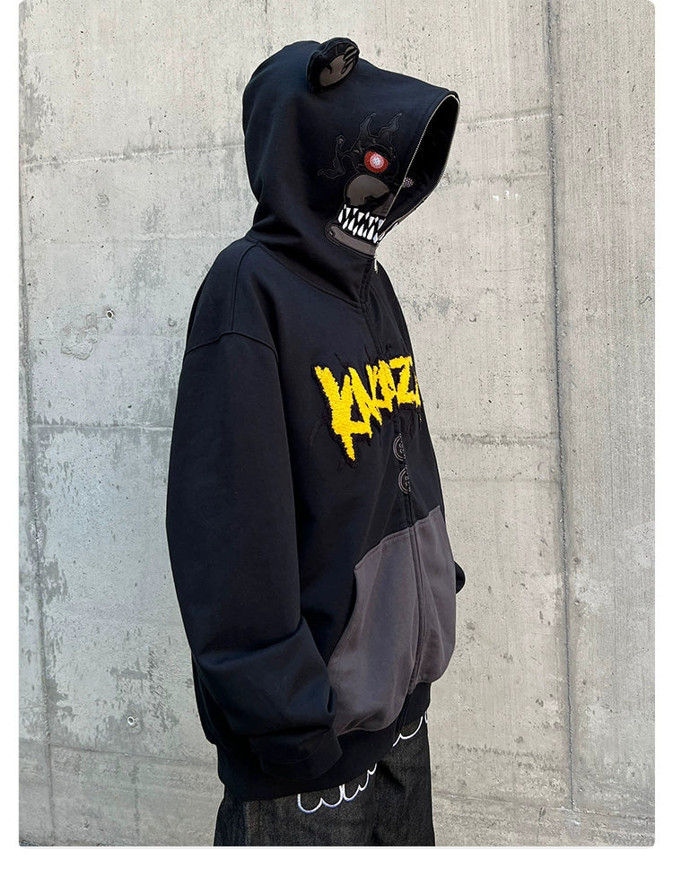 A horror fan wears the Seakoff Dark Bear Demon Hoodie, crafted from 400GSM cotton. This black hoodie features bear ears, a stylized yellow logo, menacing red eyes and sharp teeth on the hood, dark pockets, and a loose fit for comfort against a concrete wall.