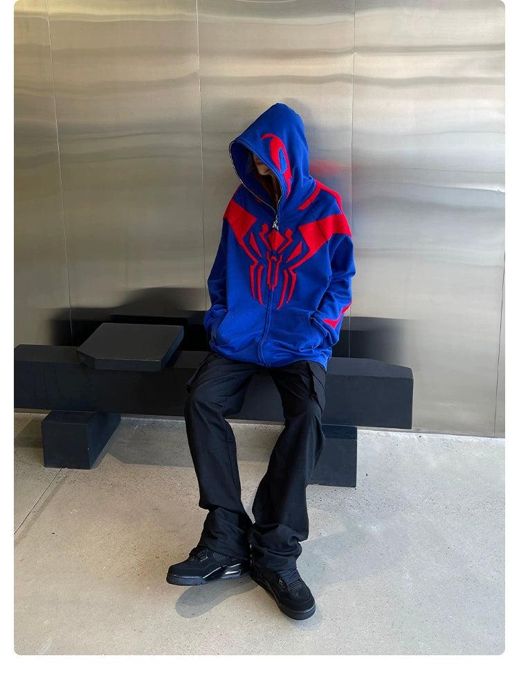 A person in a Seakoff Superhero Spider Hoodie, a 400GSM oversized blue and red zip-up sweatshirt, sits on a black bench wearing black pants. With the hood up, their face is obscured against metallic walls and a concrete floor, enhancing the streetwear vibe.