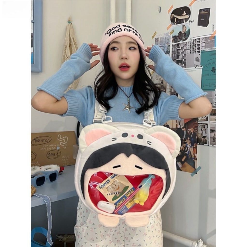 A person in a pink beanie, blue shirt, and white overalls stands in a photo-decorated room holding the Seakoff Adorable Cat Hoodie Plush Bag—an original design featuring a transparent mouth pocket.