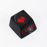 A black keyboard keycap from the Naruto Elements Keycap Set features a red heart design on top and "LOVE" in red on the side. It has a glossy finish with subtle speckles, blending charm with Naruto&
