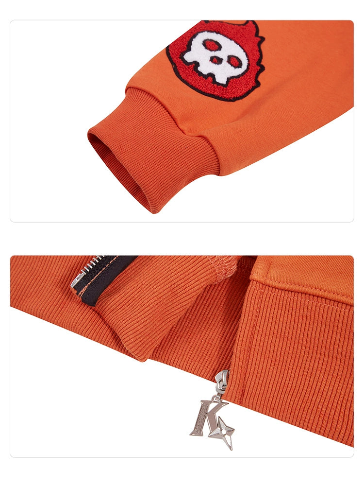 A close-up of the Fire Force Inspired Orange Zip-Up Hoodie by Seakoff shows a sleeve adorned with a red skull patch, defining streetwear fashion. Below, the ribbed hem features a silver zipper and charm, made from 400GSM knit fabric with a ribbed texture.